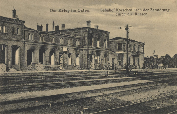 Schwarzes und weißes Foto einer Bahnstation in Korschen Ost, Deutschland, mit Schienen, Gebäuden, Strommasten, Menschen, Bäumen und Himmel.