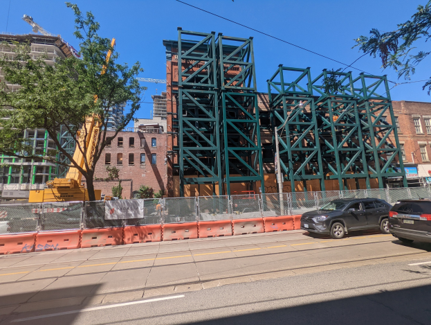 Große Metallbrücke im Bau an einer Stelle mit parkenden Autos, einem Zaun, Bäumen, Gebäuden, einem Kran und Himmel im Hintergrund.