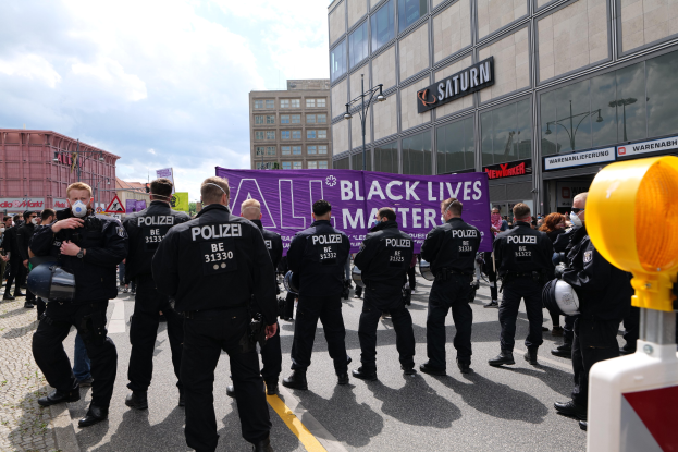Police officers in uniform stand facing a crowd, some of whom wear helmets and hold firearms, with a banner, buildings, light poles, and text boards in the background under a cloudy sky and a traffic signal on the right.