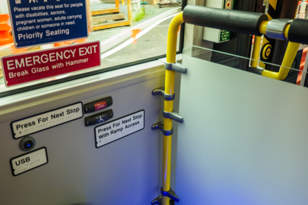 Notausgangsschild an einer Zugseite, mit Türgriff im Vordergrund, Glasfenster im Hintergrund, das Straße, Pfosten und externe Objekte zeigt.