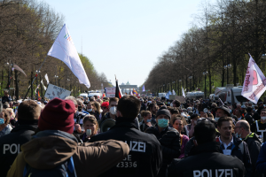 Eine große Gruppe von Menschen steht vor einer Reihe von Polizeibeamten, einige tragen Mützen und Masken, während sie bei einer Demonstration in Berlin, Deutschland, mit Schildern, Fahnen, Laternenpfählen, Bäumen, Fahrzeugen, einem Gebäude und dem Himmel im Hintergrund demonstrieren.