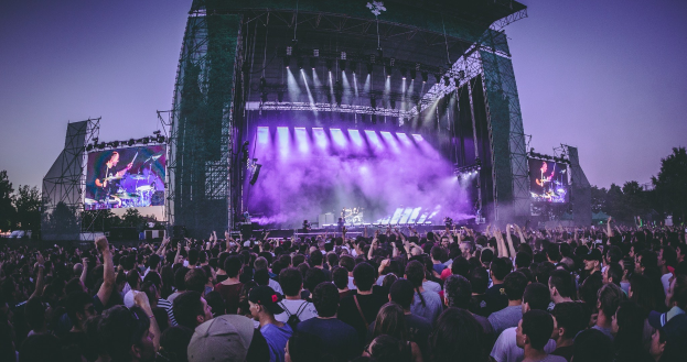 Eine große Menschenmenge bei einem Musikfestival schaut auf eine beleuchtete Bühne mit Instrumenten und Bildschirmen, die vor einem Gebäude, Bäumen und einem blauen Himmel steht.