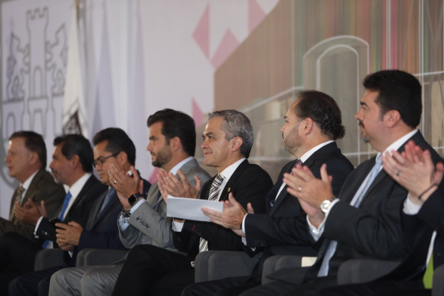 A group of men in suits and ties, some wearing glasses, sitting on a stage and clapping, with a plain wall in the background.