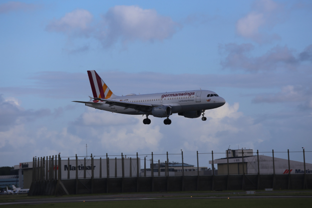 Germanwings Airbus A320-200 beim Start vom Frankfurter Flughafen, mit Gras darunter, einem Zaun im Vordergrund, Gebäuden und Bäumen im Hintergrund und einer bewölkten Himmel