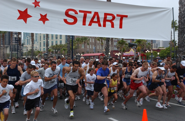 Gruppe von Läufern bei einem Marathon, die an einer Verkehrsinsel vorbeilaufen, mit einem Banner und städtischer Umgebung unter einem klaren Himmel.