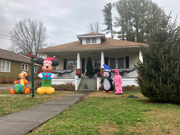 A house with windows, pillars, railings, steps, and a roof, surrounded by potted plants, grass, a pathway, a group of trees, wires, and a cloudy sky, featuring Mickey Mouse and Minnie Mouse Christmas decorations in front.