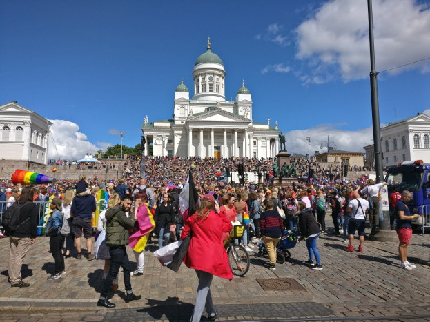 A large crowd walks down a street in front of a building with windows, pillars, and arches, some holding flags and pushing strollers, with parked vehicles, light poles, trees, and a cloudy sky in the background.