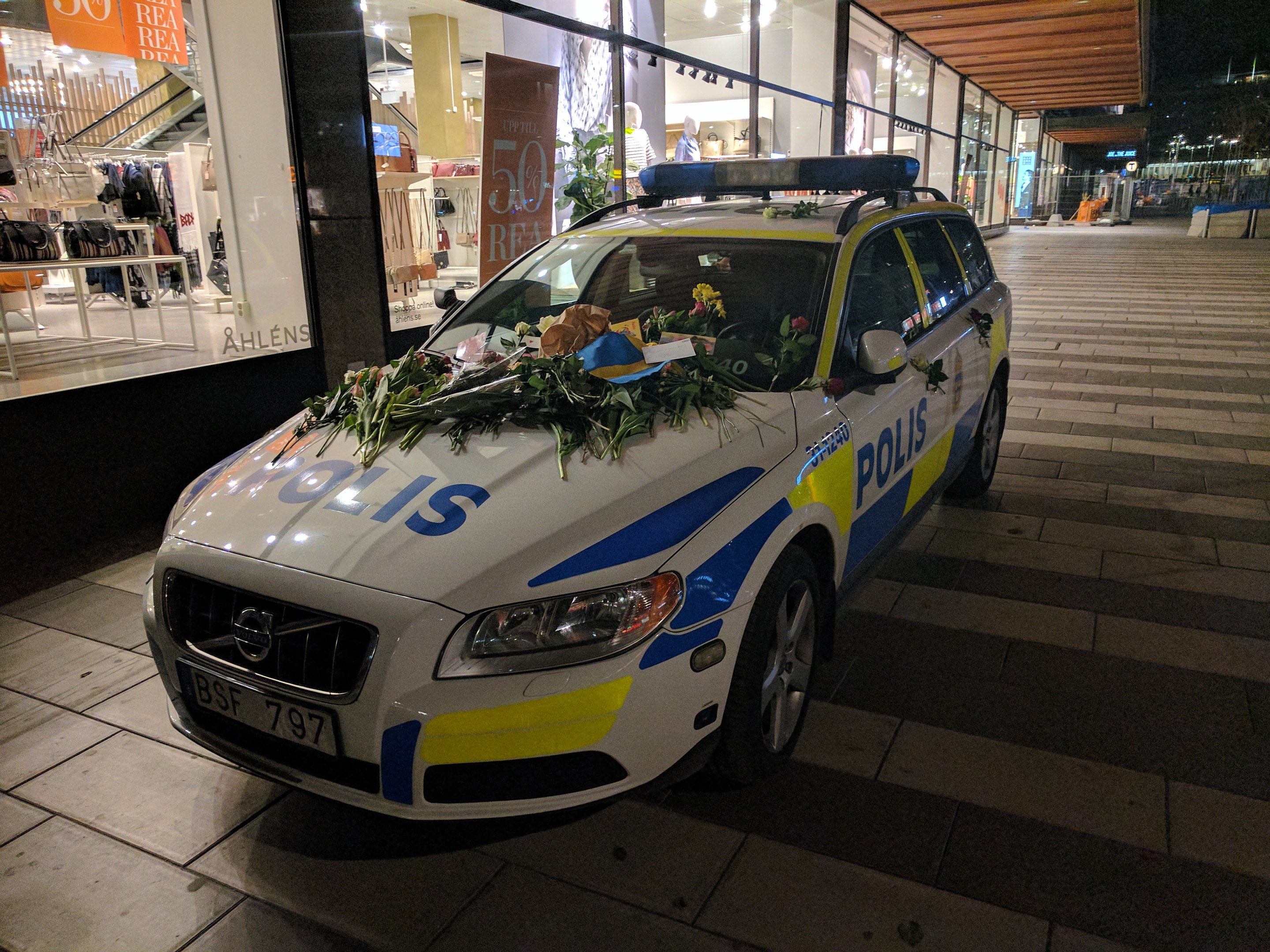 Ein mit Blumen und Blättern geschmückter Polizeiwagen steht nachts vor einem Geschäft mit Glaswänden, das Schaufensterpuppen in Kleidern und beleuchtete Schilder zeigt.