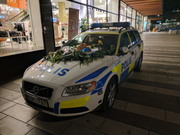 Ein mit Blumen und Blättern geschmückter Polizeiwagen steht nachts vor einem Geschäft mit Glaswänden, das Schaufensterpuppen in Kleidern und beleuchtete Schilder zeigt.