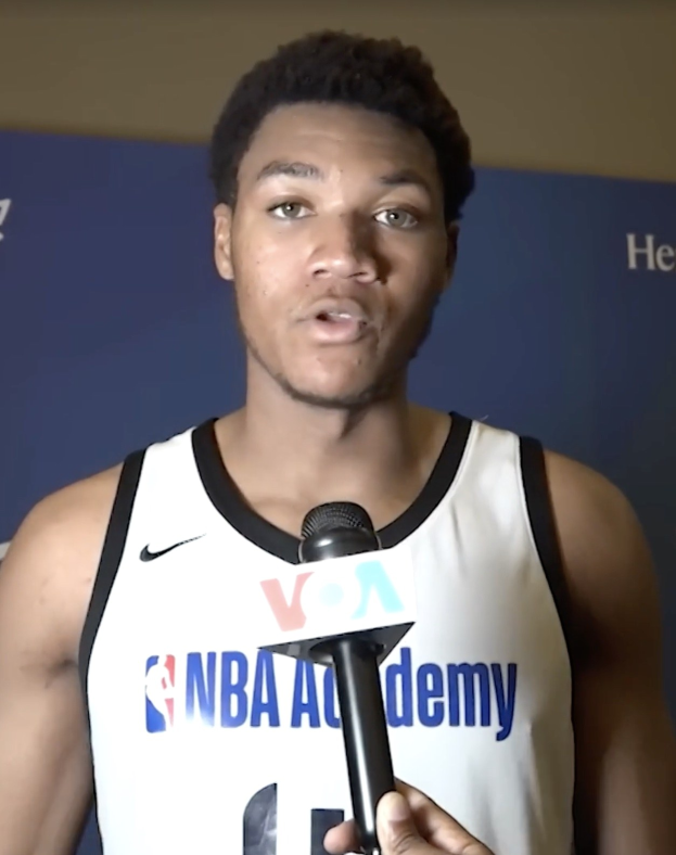 Ein Mann in einem weißen Tanktop hält ein Mikrofon und spricht vor einem Banner "NBA All-Star Game 2019" mit der Presse.