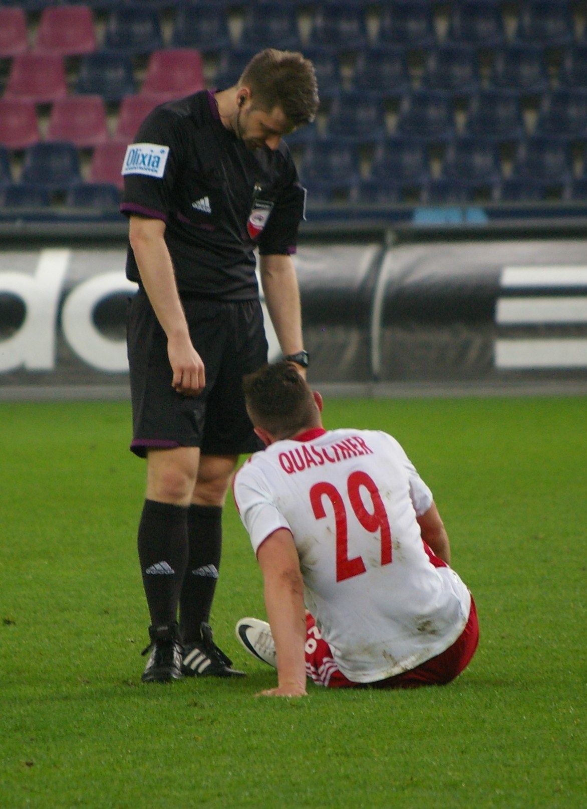 Ein Fussballspieler in Sportkleidung sitzt neben einem Schiedsrichter auf dem Boden in einem Stadion.