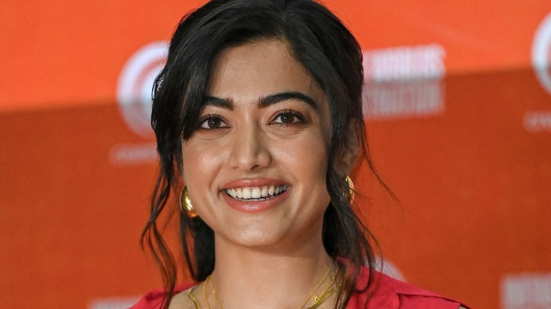 Eine Frau in einem leuchtend roten Kleid mit einem eleganten Dutt und dezentem Make-up posiert strahlend vor der Kamera, trägt schwarze High Heels und hat ein hellgelbes Banner mit der Aufschrift 'Die besten Schönheits Looks von Sonam Kapoor beim Cannes Film Festival' im Hintergrund.