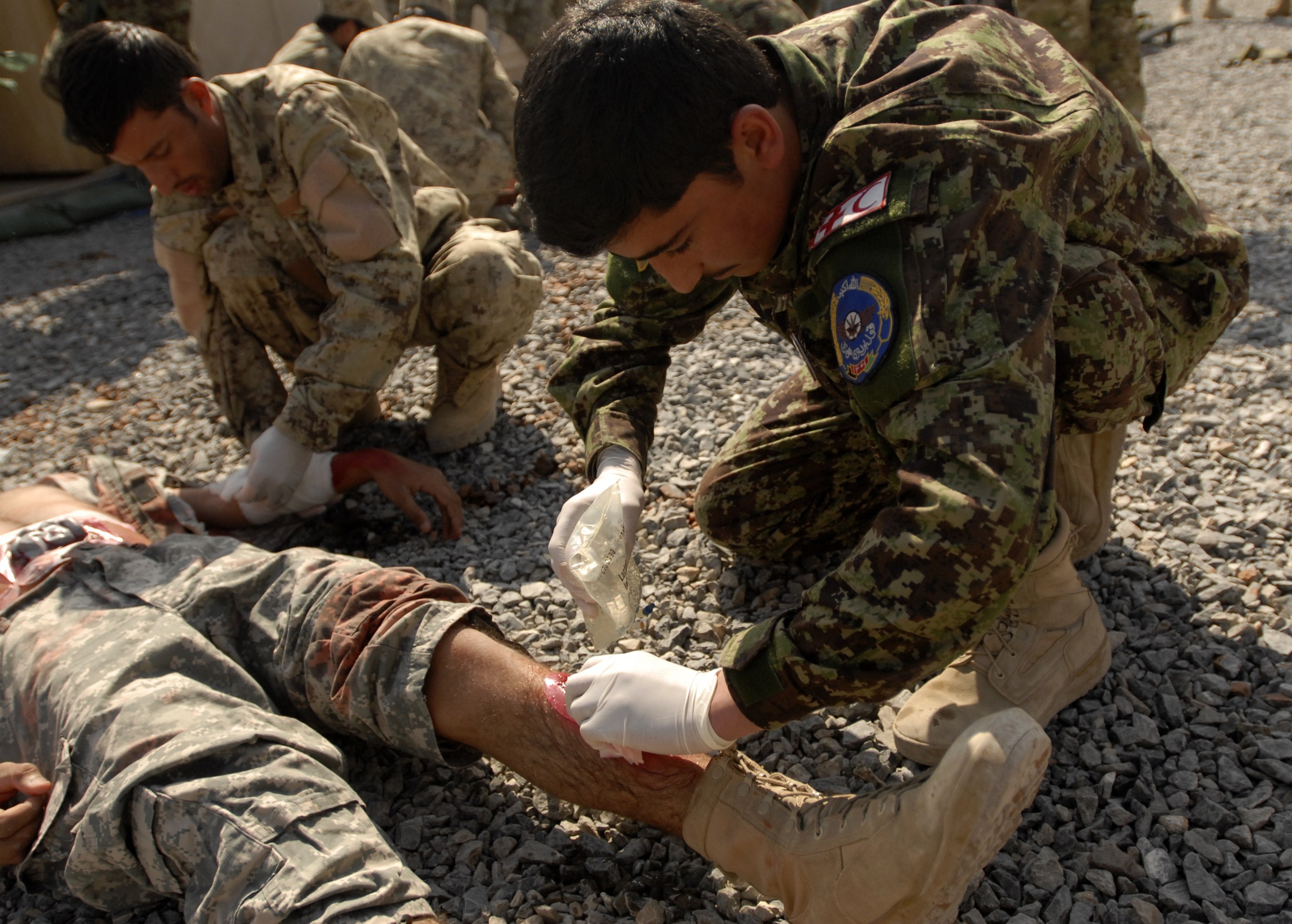 A group of men in military uniforms kneeling on the ground, with one lying on stones, one wearing a bandage on his leg; a tent is visible on the left and others stand in the background.