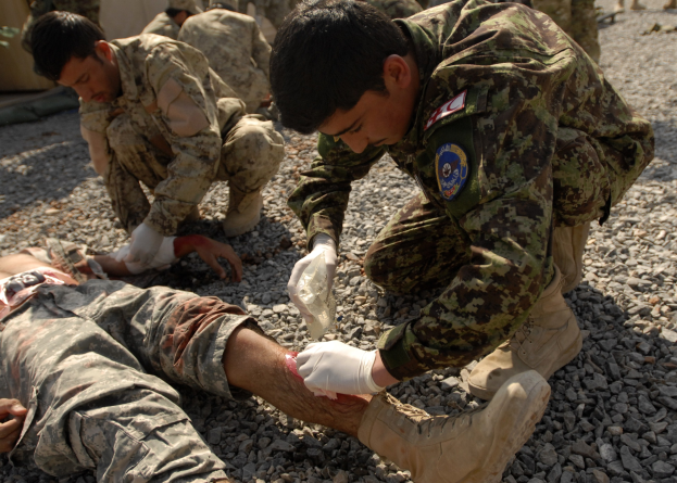 A group of men in military uniforms kneeling on the ground, with one lying on stones, one wearing a bandage on his leg; a tent is visible on the left and others stand in the background.