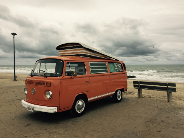 Ein oranger VW-Bus auf einem Strandabschnitt neben dem Meer geparkt, mit einer Bank und einem Laternenmast in der N├Ąhe unter einem bew├Âlktem Himmel.