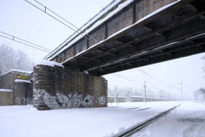 Eine schneebedeckte Bahnstrecke unter einer Brücke mit Graffiti, Strommästen mit Drähten, Bäumen und einem Himmel im Hintergrund.