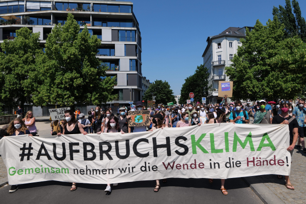 A group of people wearing masks hold a banner reading "Aufbruchsklima" in front of a building with trees and a clear sky behind them.