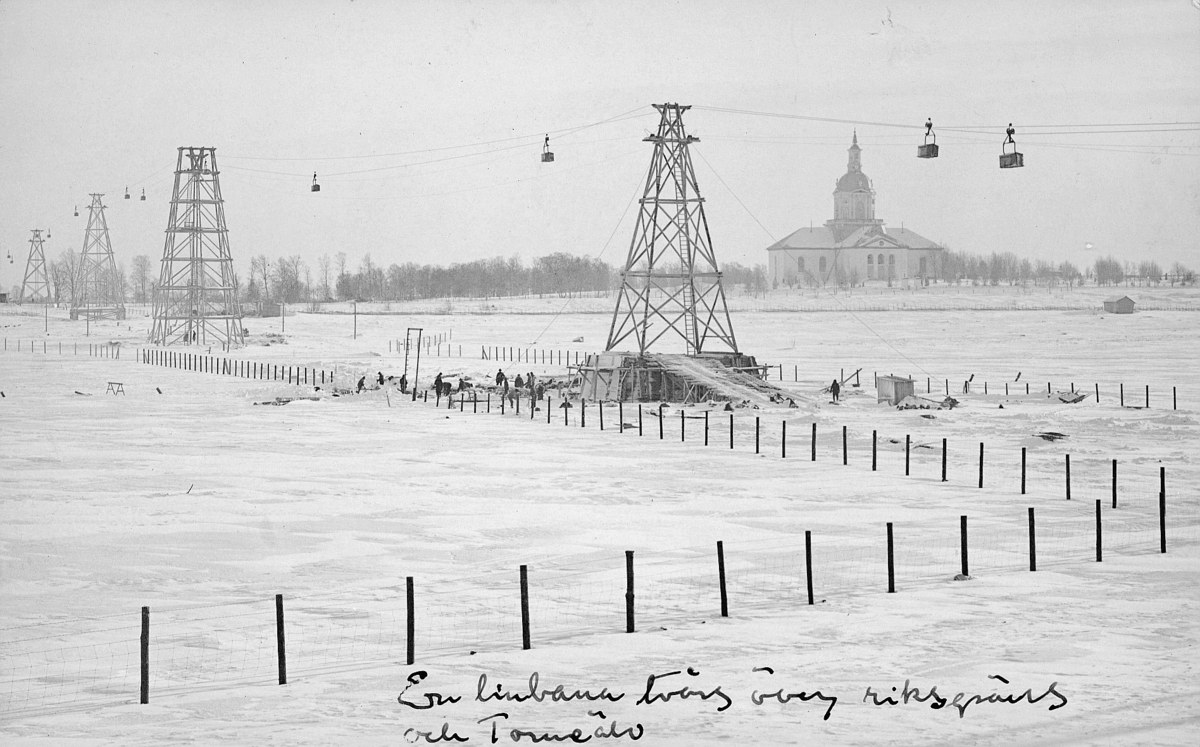 Schwarze-Weiß-Foto eines Skilifts in einem verschneiten Feld mit Stützpfählen, Überseilbahn, Bäumen und einem Gebäude im Hintergrund, mit Text am unteren Rand.
