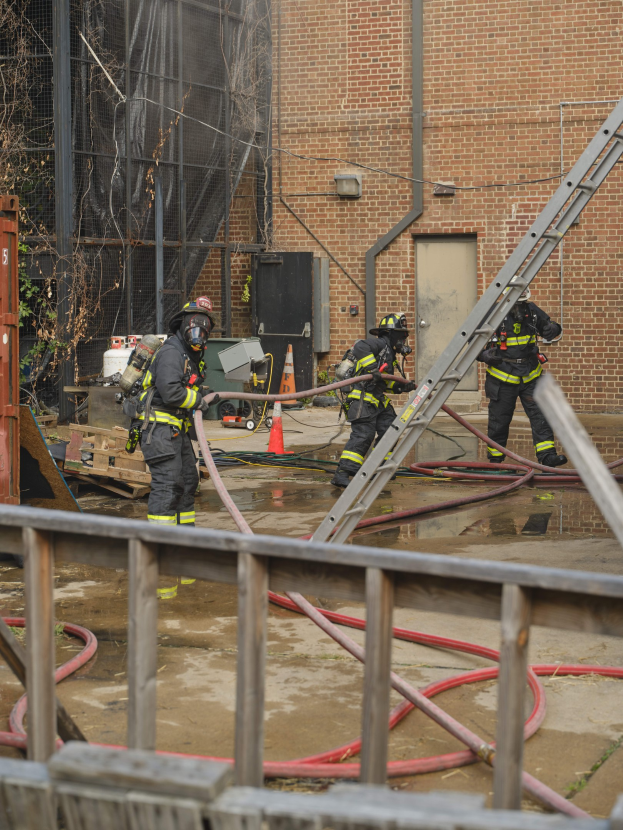 Feuerwehrleute in Helmen arbeiten daran, ein Gebäude Feuer zu löschen, umgeben von Ausrüstung und einem Metallzaun.