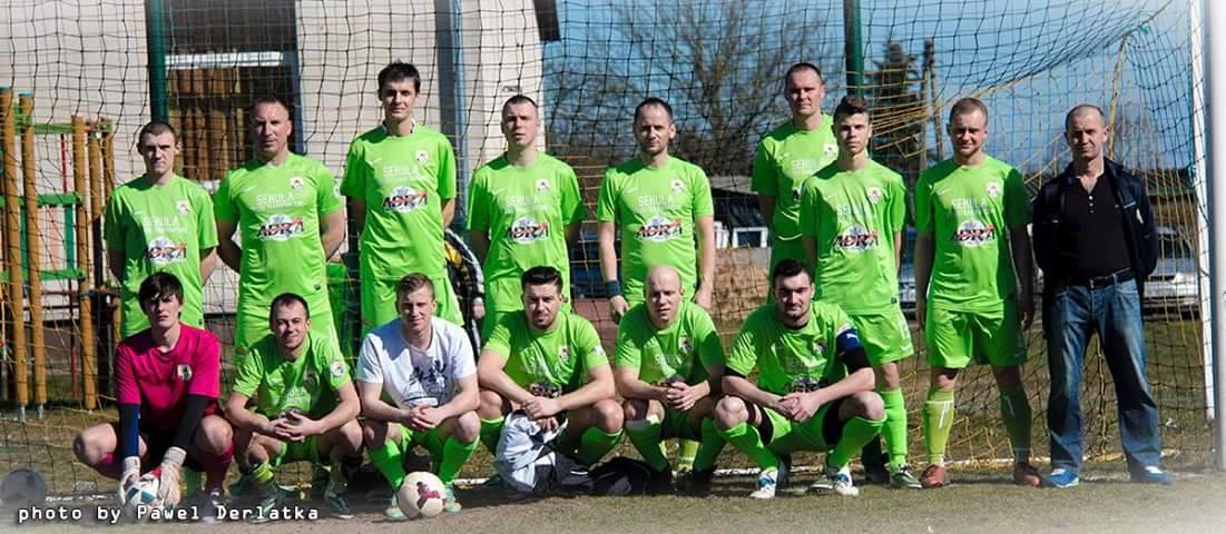 Eine Gruppe von Männern in grünen T-Shirts steht auf einem Fußballfeld, einige in Hockstellung, mit einem Ball vor ihnen in der Nähe eines Torpfostens, Bäume, Gebäude und Fahrzeuge im Hintergrund.