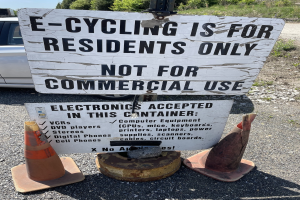 Schild mit der Aufschrift "E-Bike-Fahren nur für Anwohner" steht an einer Straße mit Verkehrskegeln in der Nähe, ein Fahrzeug im Hintergrund, Bäume und ein klarer blauer Himmel.