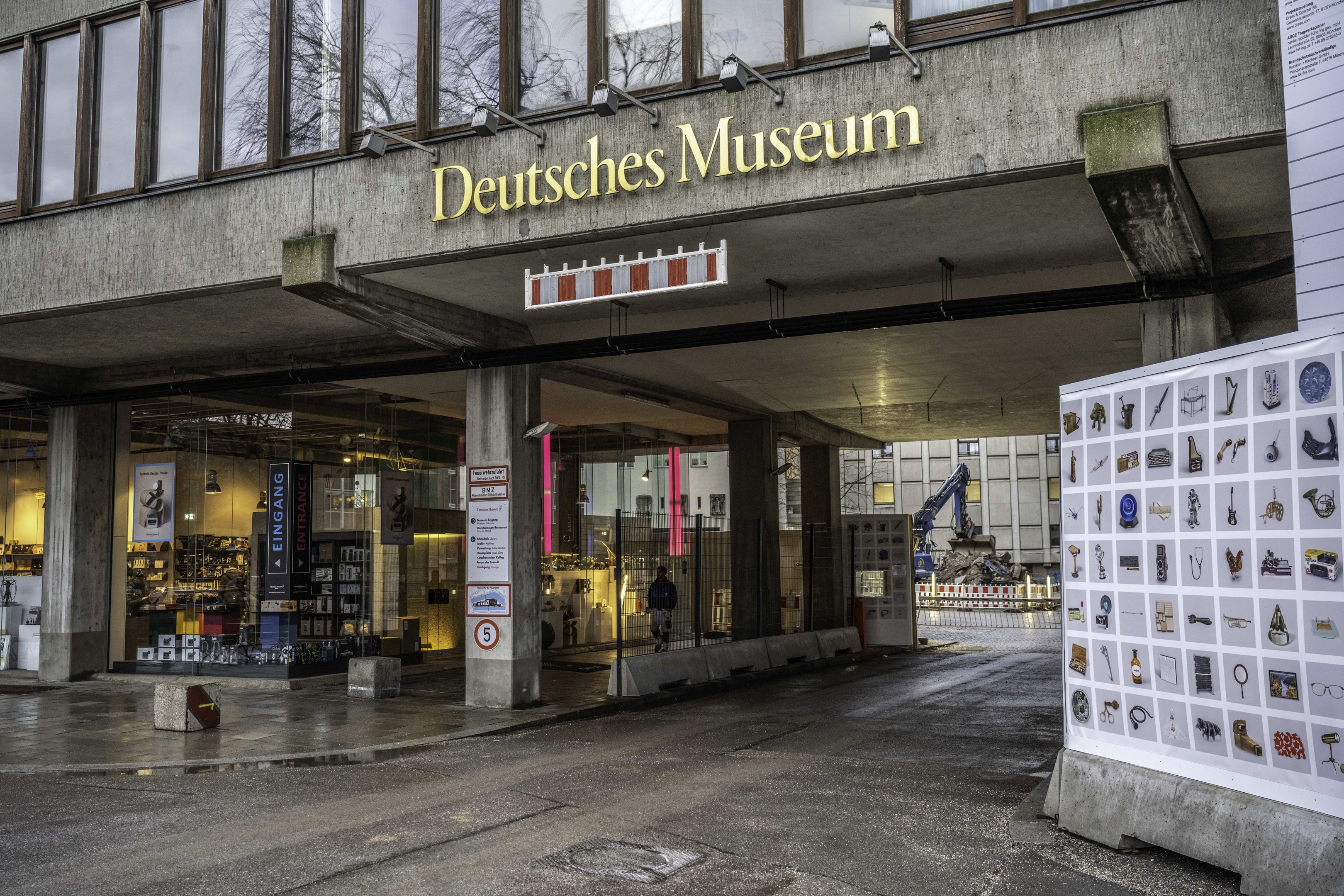Großes gläsernes Gebäude des Deutschen Museums in Berlin mit einem Namensschild und einer Informationsanzeige rechts daneben, vor einem Hintergrund aus anderen Gebäuden, Bäumen und Himmel.