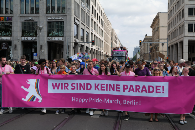 Eine Gruppe von Menschen marschiert auf einer Berliner Straße mit einem pinken Banner, auf dem 'Happy Pride March' steht, vorbei an Gebäuden, Laternenpfählen und Verkehrszeichen unter einem bewölkten Himmel.