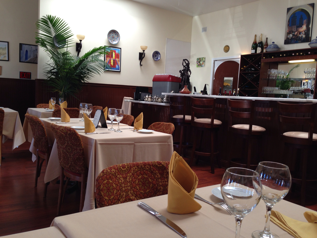 Ein Restaurantinnenraum mit Tischen und Stühlen, einem langen Tisch mit Gläsern, Servietten, Tellern und Löffeln, einem Regal mit Flaschen und Gläsern, einer Wand mit Fotorahmen, einer Uhr, zwei Lampen und einer Pflanze vor der Wand.