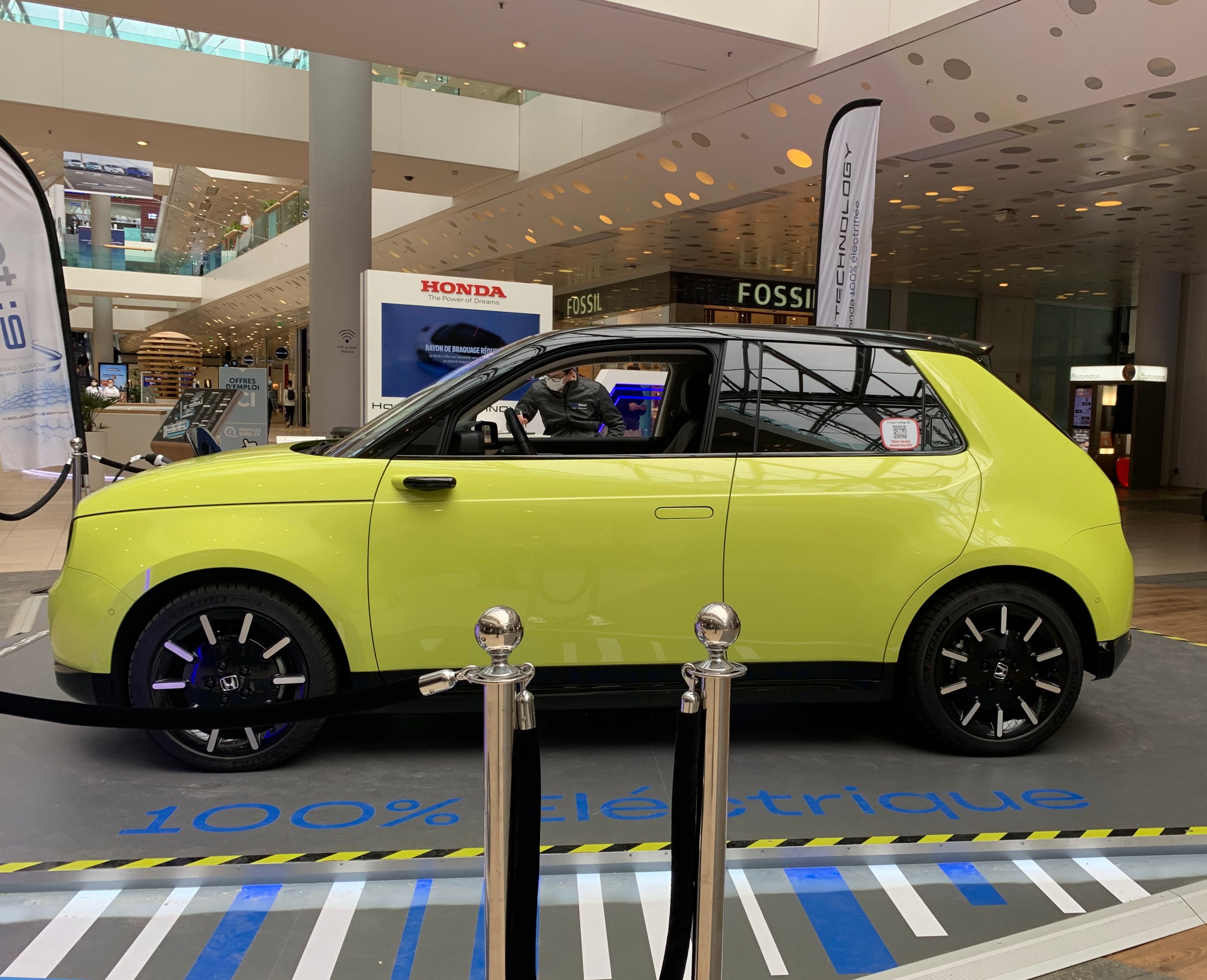 Gelber wasserstoffbetriebenes Elektroauto vor einem Gebäude mit Absperrpollern, Bannern, Menschen, Schildern, einer Topfpflanze, Deckenleuchten und Lüfter.