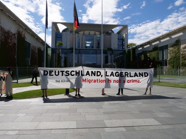 Eine Gruppe von Menschen hält ein Transparent mit der Aufschrift 'Deutschland, Lagerland Migration ist kein Verbrechen' vor einem eingezäunten Gebäude mit Bäumen und Fahnen.