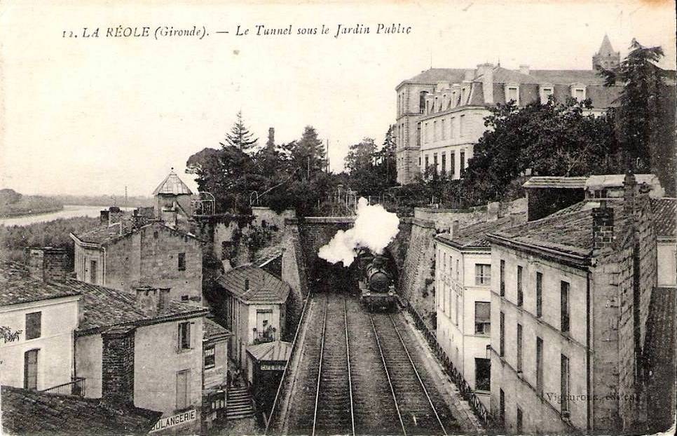 Black and white photograph of a train on tracks passing through a town, with buildings, trees, and water visible, and text at the top reading "la réole gironde - le tunnel sous le jardin public".