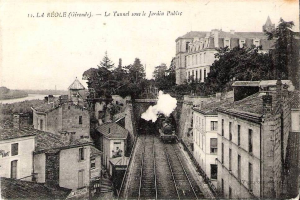 Black and white photograph of a train on tracks passing through a town, with buildings, trees, and water visible, and text at the top reading "la réole gironde - le tunnel sous le jardin public".