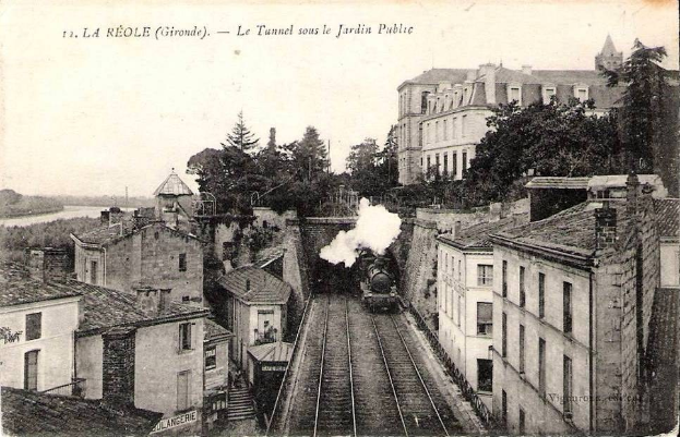 Black and white photograph of a train on tracks passing through a town, with buildings, trees, and water visible, and text at the top reading "la réole gironde - le tunnel sous le jardin public".