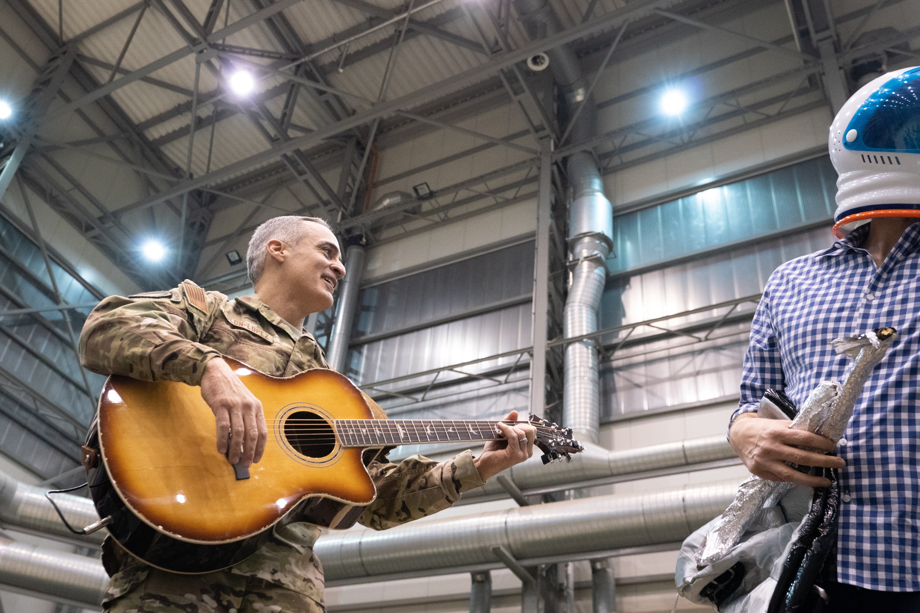 Ein Mann in Militäruniform spielt eine Gitarre, waehrend er neben einem anderen Mann steht, der einen Helm trägt, mit Rohren und Lichtern im Hintergrund.