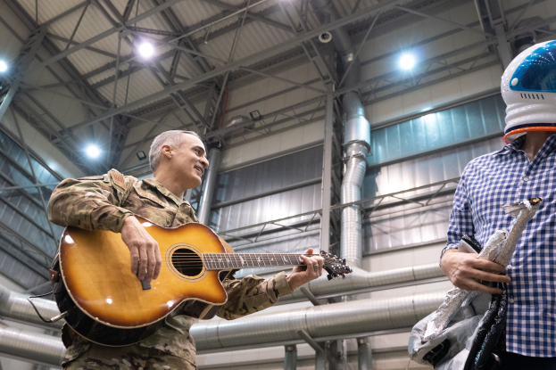 Ein Mann in Militäruniform spielt eine Gitarre, waehrend er neben einem anderen Mann steht, der einen Helm trägt, mit Rohren und Lichtern im Hintergrund.