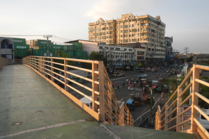 Fußgängerbrücke mit Geländern, die eine vielbefahrene Straße überspannt, mit Autos und Fußgängern, sowie Gebäuden, Bäumen, Strommasten mit Drähten und einem bewölkten Himmel im Hintergrund.