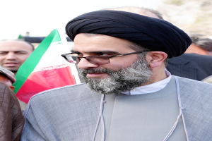 Ayatollah Ali Khamenei, der Oberste Führer des Iran, trägt ein graues Gewand und eine schwarze Kopfbedeckung mit Brillengläsern und steht vor einer Gruppe von Menschen und einer Flagge.