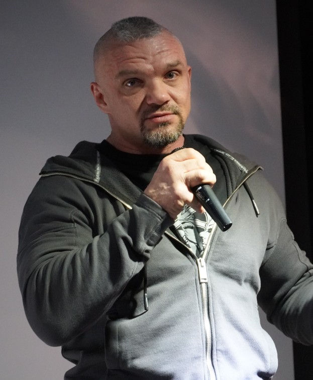 Ein Mann in einer schwarzen Kapuzenjacke und einem schwarzen Anzug mit einem weißen Hemd hält ein Mikrofon und steht selbstbewusst vor einer Wand, wobei er wie ein Komiker aussieht.