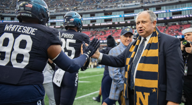 Ein Mann im Anzug und Krawatte gibt einem Fußballspieler die Hand auf einem Feld, umgeben von einer Gruppe von Menschen, während eine Person mit einer Kamera in der Nähe steht und ein Stadion mit Zuschauern und Schildern mit der Aufschrift "Notre Dame Football Coach Mike McCarthy und die Fighting Irish" im Hintergrund zu sehen ist.
