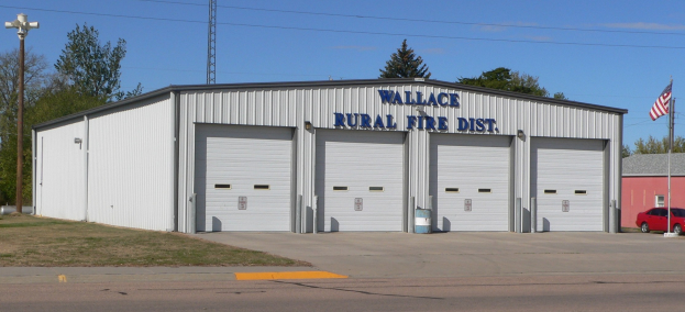 Eine Feuerwache des Wallace Rural Fire District mit einer Straße im Vordergrund, Gras auf der linken Seite, einem Lichtmast auf der rechten Seite, Bäumen im Hintergrund, einem Turm in der Mitte und einer Flagge, die im Wind weht, unter einem sichtbaren Himmel.