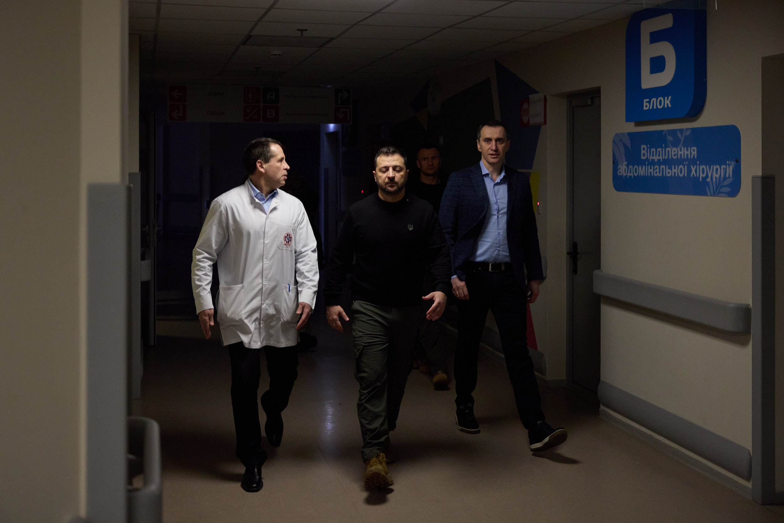 Eine Gruppe von Männern geht einen Krankenhausflur entlang, der von Deckenlampen beleuchtet wird, mit einer Tür auf der rechten Seite und Texttafeln auf der linken Seite, die besorgt aussehen.