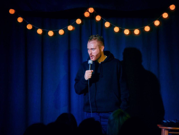 A man standing on a brightly lit stage holding a microphone, with a table and curtain in the background, addressing an audience at a stand-up comedy event.