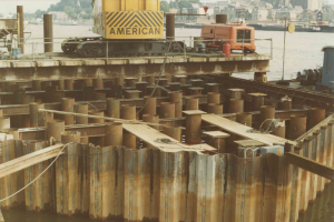 Baustelle mit einem Kran, der Stahlstäbe aus einem Stapel hebt, umgeben von Wasser, Gebäuden, Bäumen und einem klaren blauen Himmel.