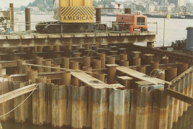 Baustelle mit einem Kran, der Stahlstäbe aus einem Stapel hebt, umgeben von Wasser, Gebäuden, Bäumen und einem klaren blauen Himmel.