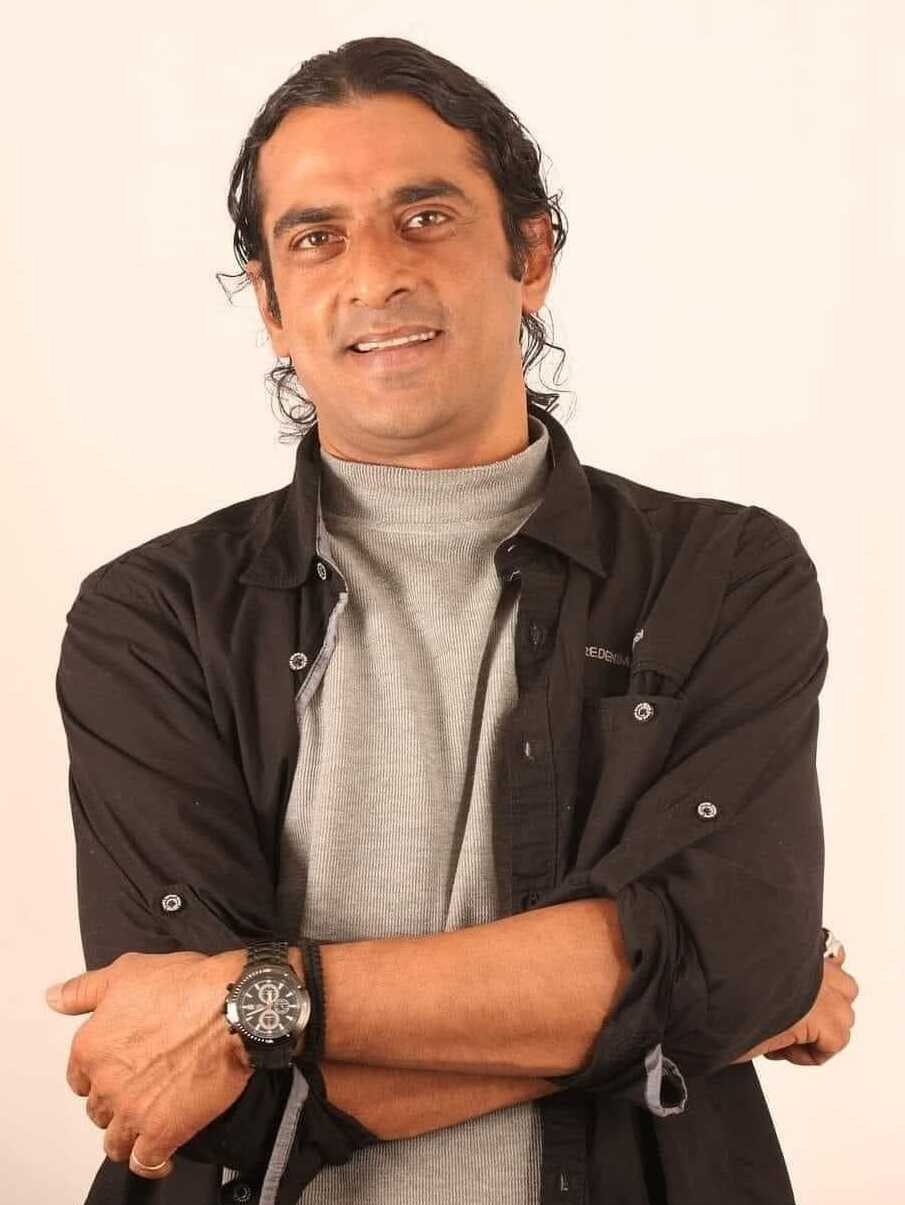 Ein Mann in einem schwarzen und grauen Outfit mit überkreuzten Armen und einer Uhr steht vor einem weißen Hintergrund und sieht nachdenklich aus.