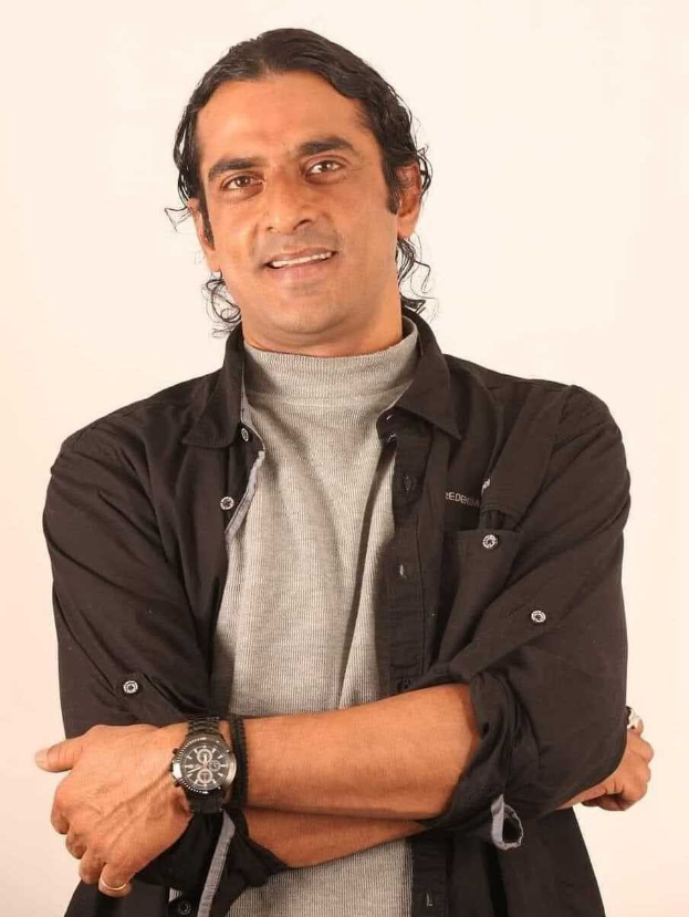 Ein Mann in einem schwarzen und grauen Outfit mit überkreuzten Armen und einer Uhr steht vor einem weißen Hintergrund und sieht nachdenklich aus.