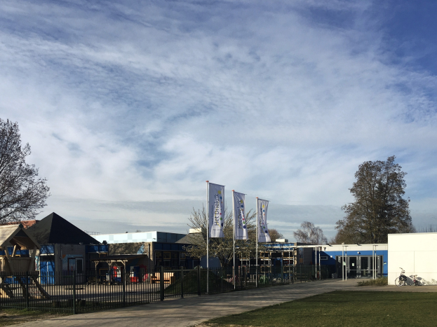 Großes Gebäude mit einem grasigen Bereich davor, umgeben von einem Metallzaun, mit einem Weg, Bäumen, Pfosten, Bannern, einem geparkten Fahrrad, nahen Häusern und einem bewölkten Himmel, als neuer Spielplatz an der Schule identifiziert.