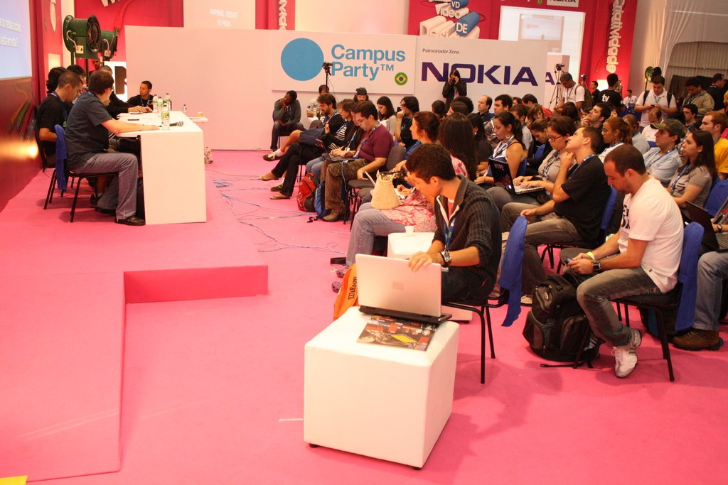 Conference with audience seated facing a stage where speakers are present, media and owners conversing, large hoarding visible in the background.