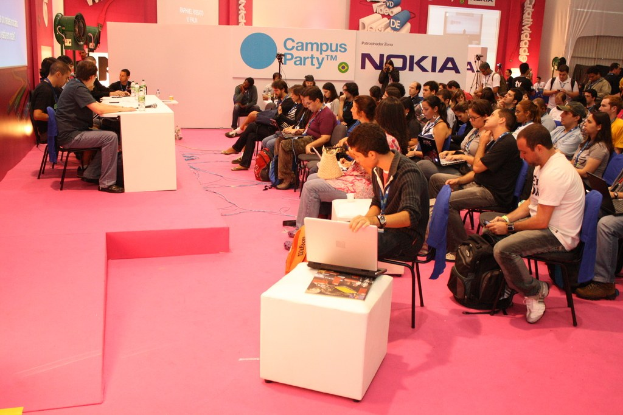 Conference with audience seated facing a stage where speakers are present, media and owners conversing, large hoarding visible in the background.