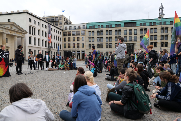Eine Gruppe von Menschen sitzt auf dem Boden vor einer Menge mit Fahnen und Transparenten während einer anti-schwulen Demonstration in Berlin, mit einem Mikrofonständer, einer Statue und Gebäuden im Hintergrund.